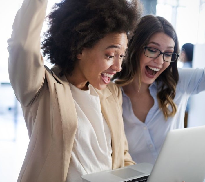 Two happy businesswomen celebrating success while reviewing HR consulting results on a laptop.